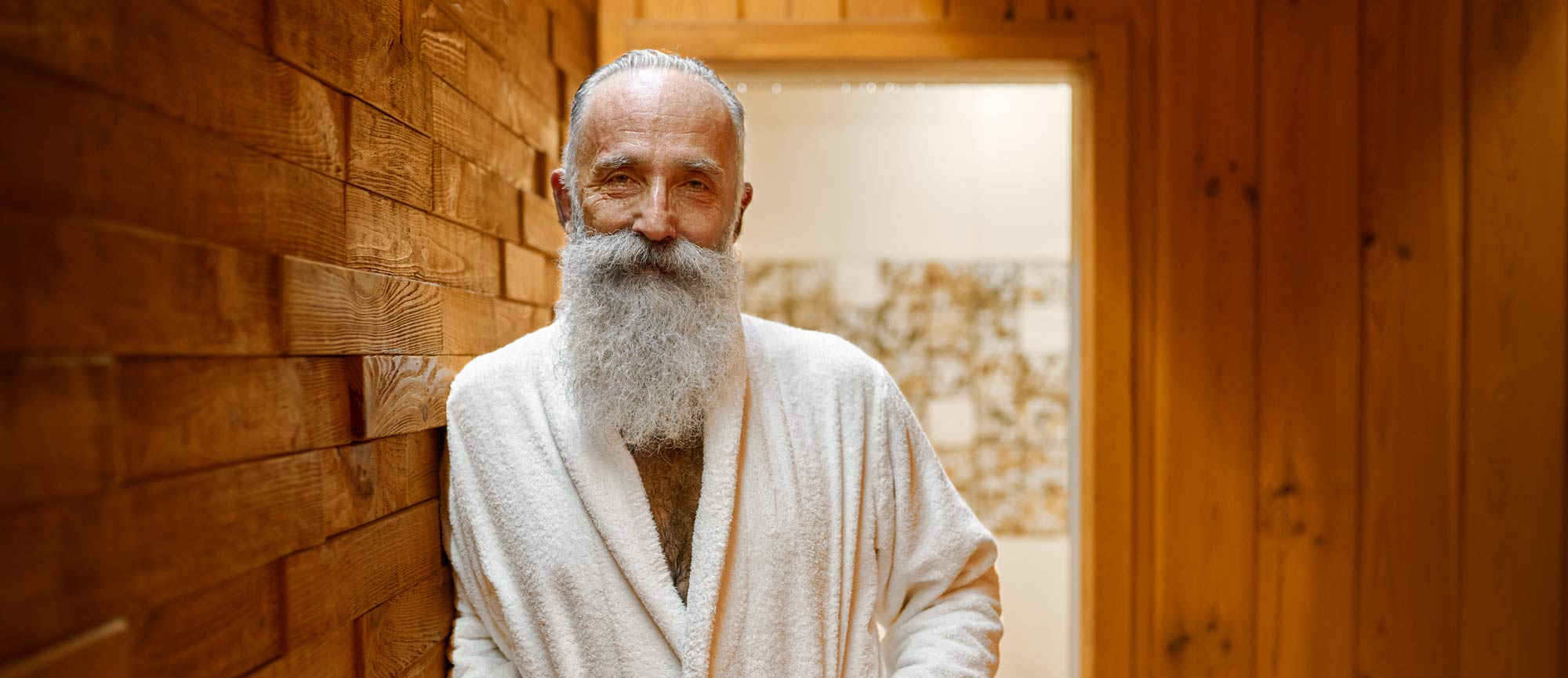 An healthy looking old man with long white beard, wearing a toweling robe and standing in a sauna, contentedly looking straight into the camera