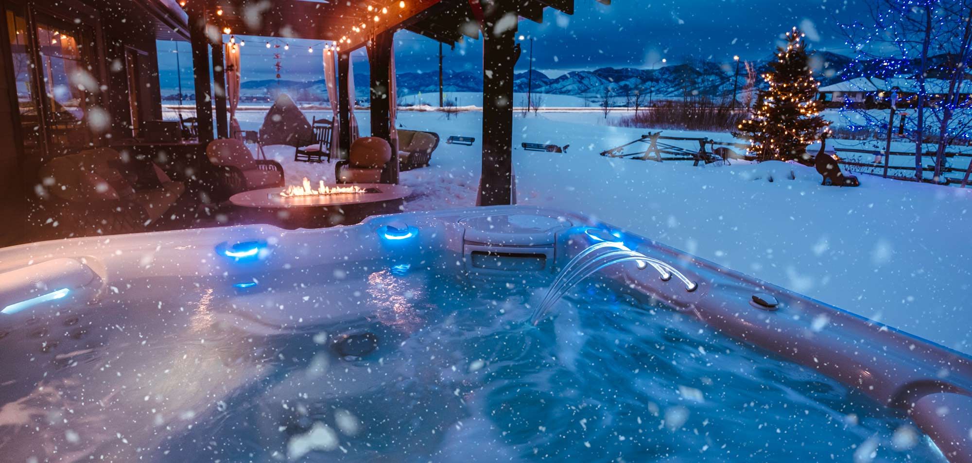 A steaming hot tub in the foreground of a winter scene showing a fairy-lit porch of what might be a wooden cabin, a christmas tree, and snow all the way to the mountains in the distance
