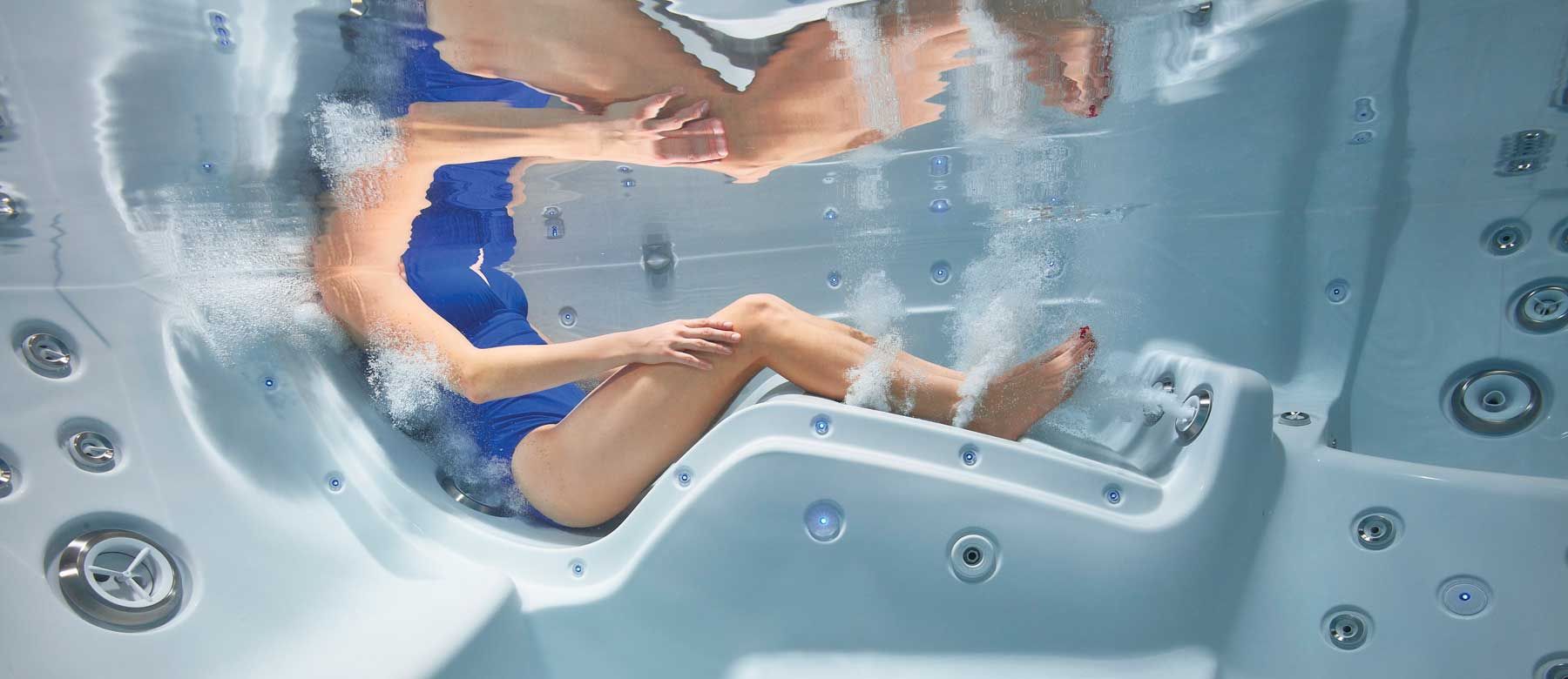 An underwater shot of a woman in a blue bathing suit sitting in a hot tub and being massaged by hydrotherapy jets