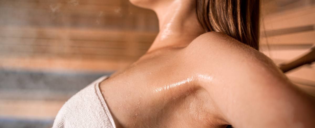 Close up on a woman sitting in a sauna wearing a towel, her shoulders, arms and neck visibly sweating